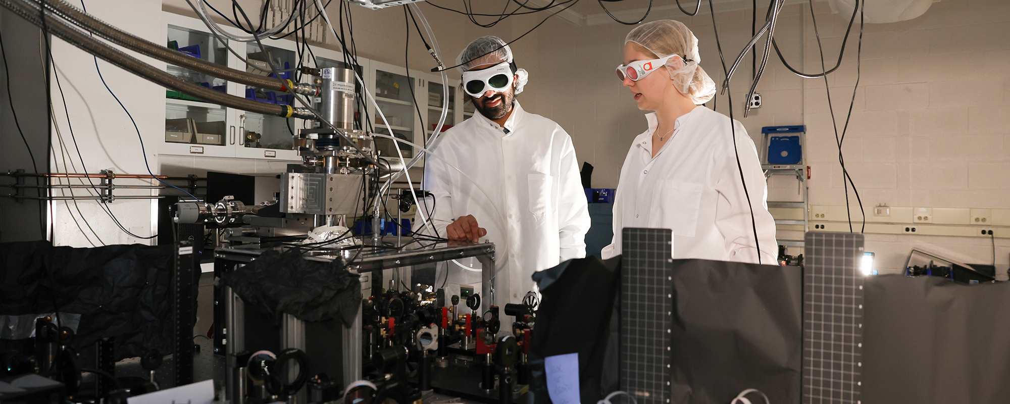 Professor Ankit Disa and an undergraduate student wear safety goggles, lab coats and hair nets in the Disa photonics lab