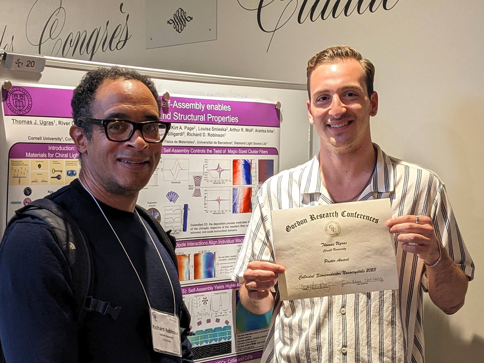Two men stand in front of a poster, one holds an award certificate.