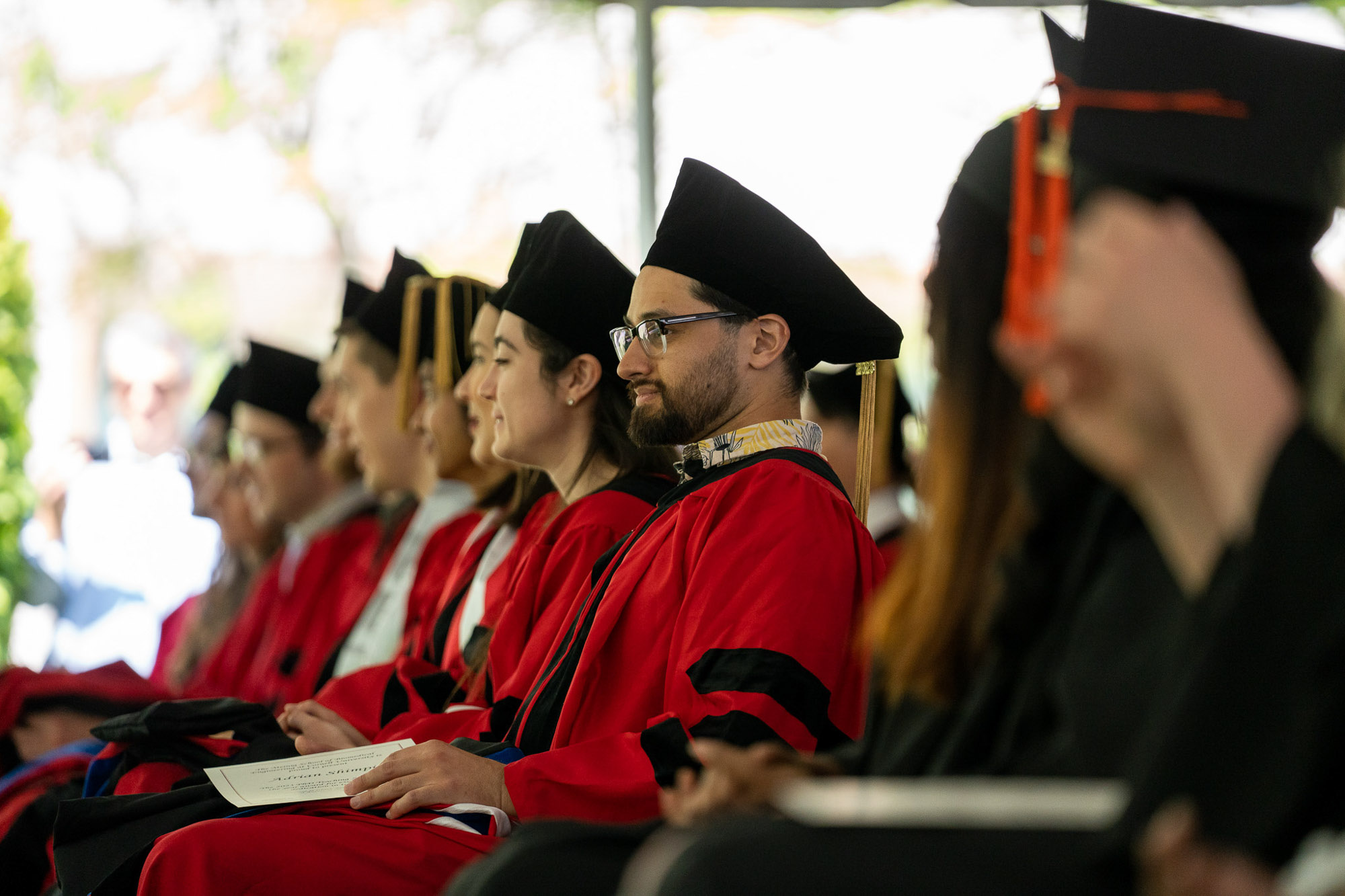 Graduate students wearing cap and gown sit with peers at graduation.
