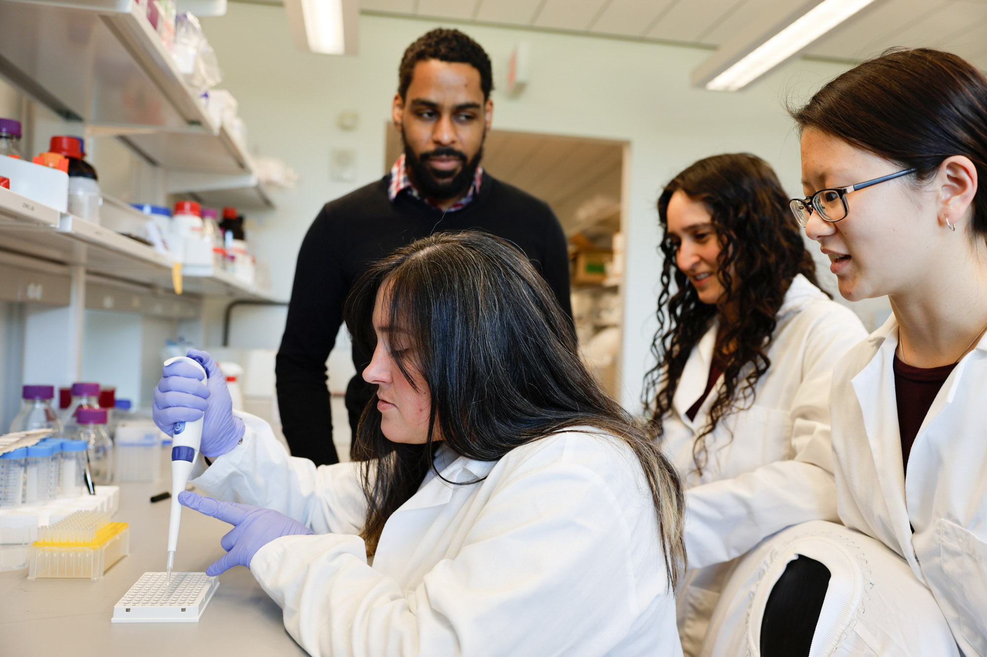 Professor Karl Lewis and two students in white lab coats watch as a third student in a white lab coat with purple gloves conducts an experiment in the Lewis lab.
