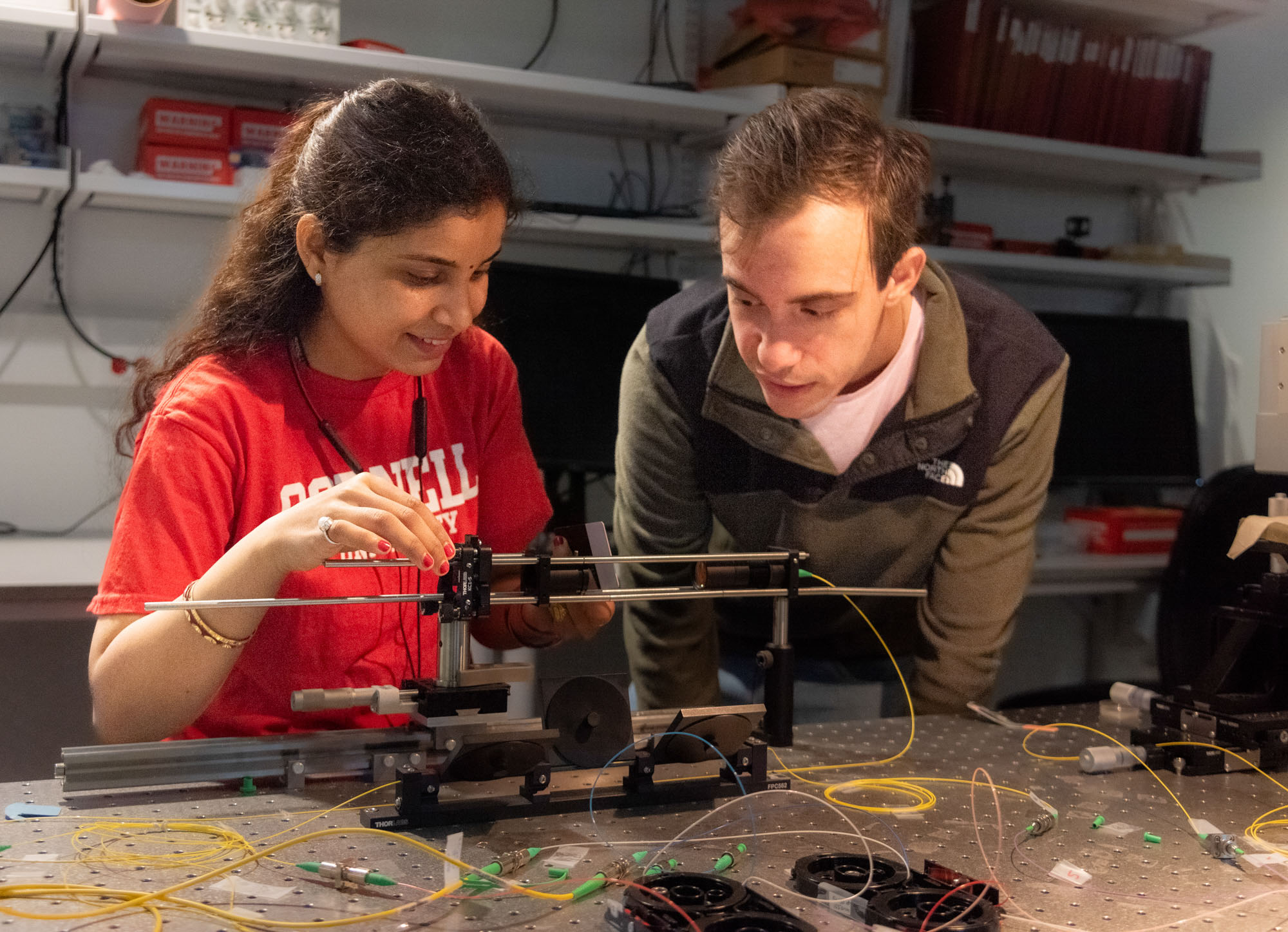 Two researchers - one wearing a red Cornell t-shirt and the other a green and black North Face pullover - conduct an experiment in the Adie lab