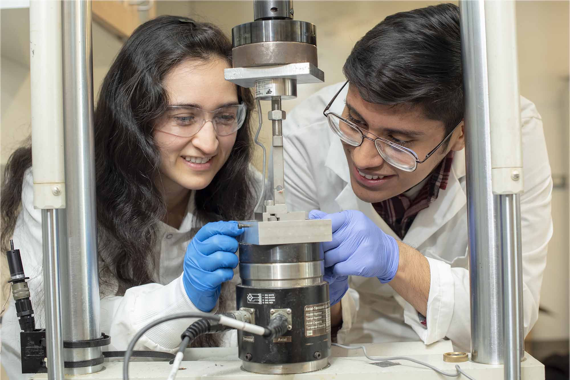 Two students in white lab coats and protective goggles examine a piece of equipment in the biomechanics lab
