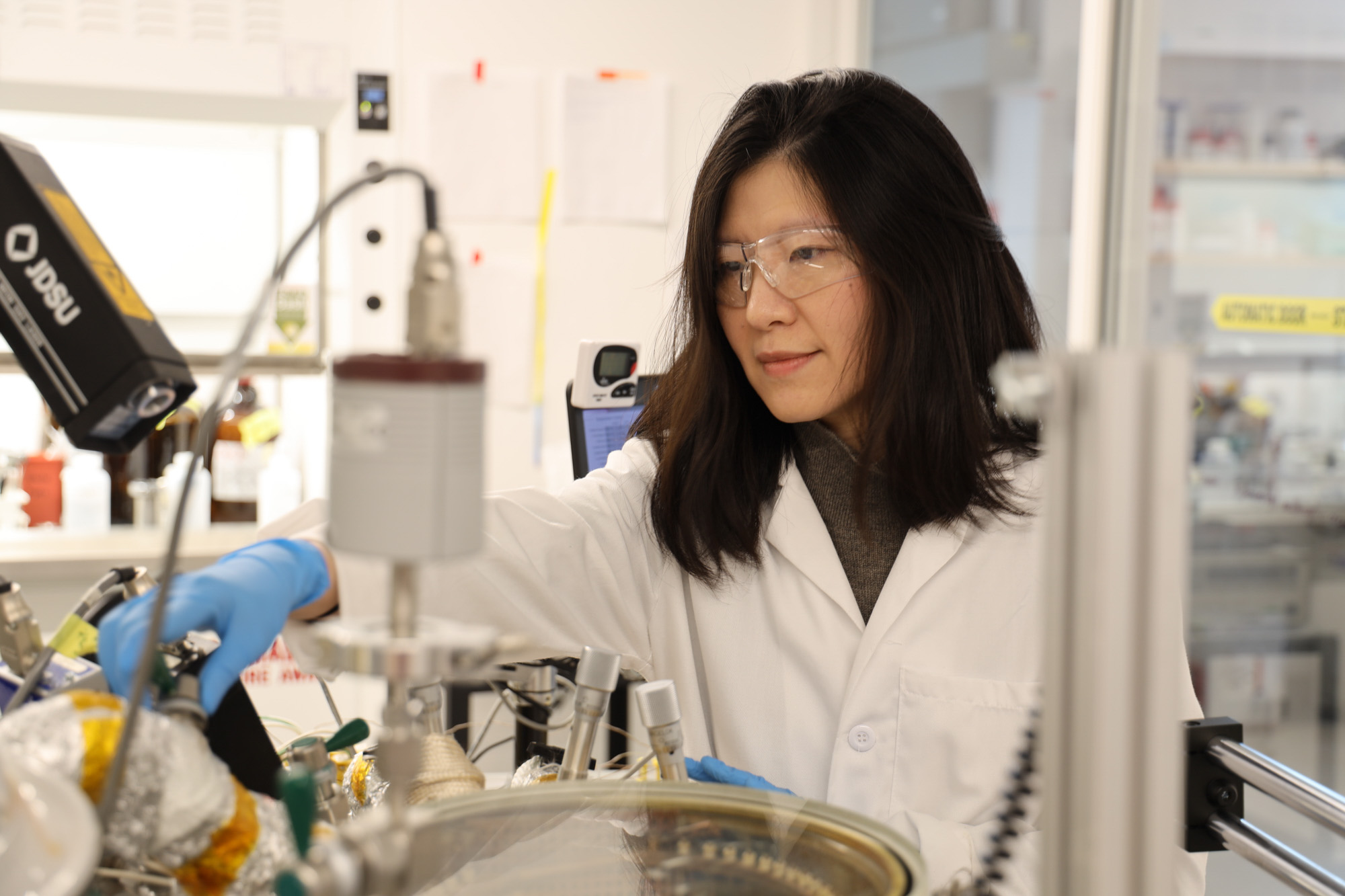 Professor Rong Yang wearing a white lab coat, safety goggles and blue gloves working in her lab