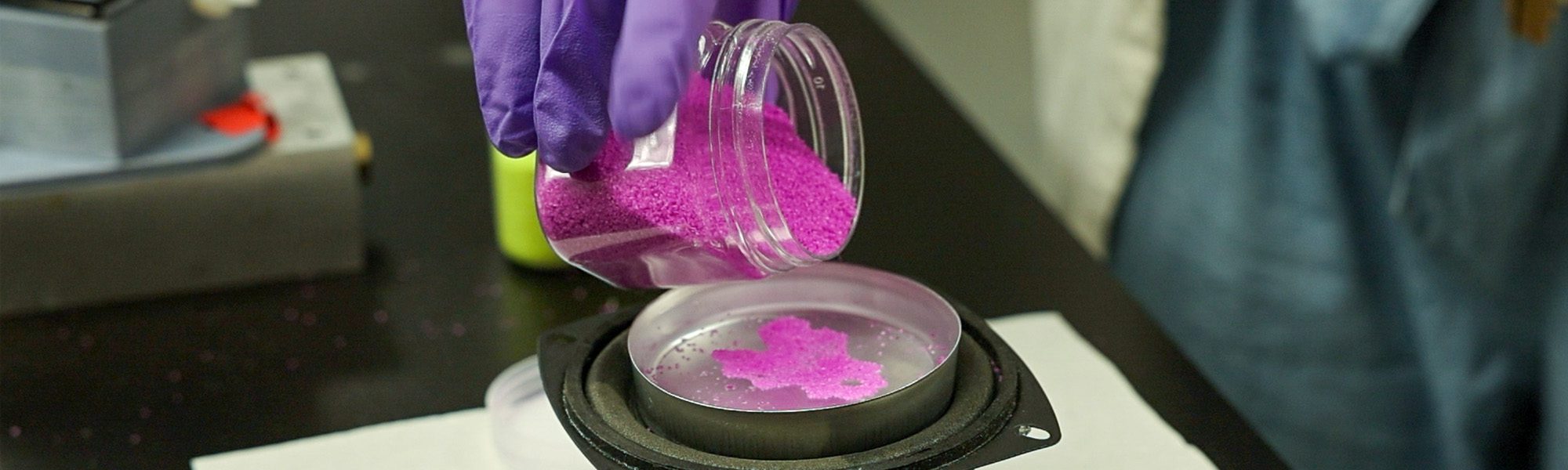 Gloved hand pours particles into liquid filled dish in the Tobias Hanrath lab.