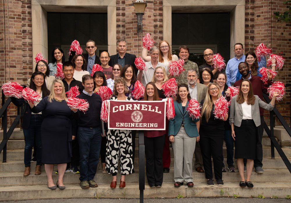 Faculty and staff from the R.F> Smith School of Chemical and Biomolecular Engineering stand with red pom poms and a Cornell Engineering banner in front of Olin Hall, a brick and stone building.