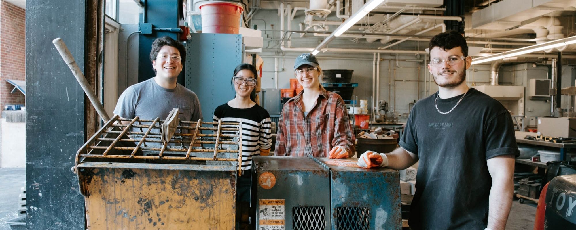 Four smiling students wearing protective glasses stand behind metal equipment