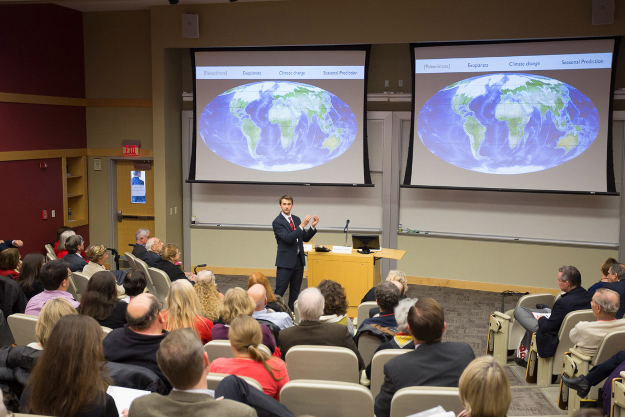 Toby Ault stands at the front of a lecture hall and gestures as he addresses a group of University trustees. There is a slide projected behind him.