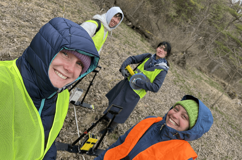 Four students in heavy winter coats smile as they take field measurements.