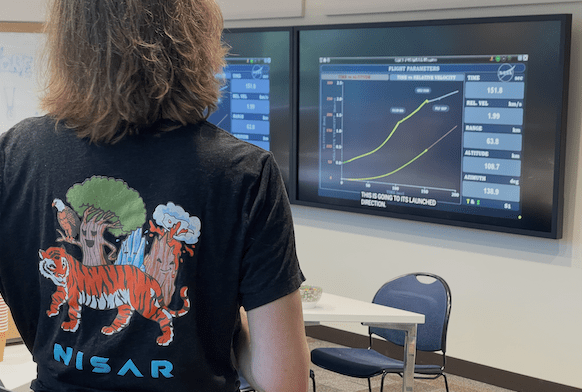 Rowena Lohman stands watching data on a screen. She is wearing a shirt with a NISAR design on the back.
