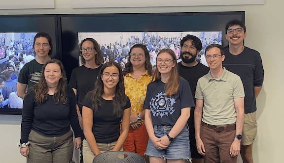 A group of nine people pose infront of two screens in Snee Hall during the launch of the NISAR satellite in 2025.