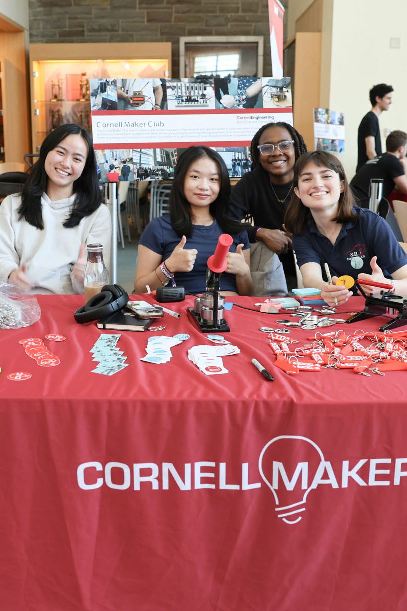 Students hold thumbs up at table that reads Cornell Maker along with stickers on and other swag items