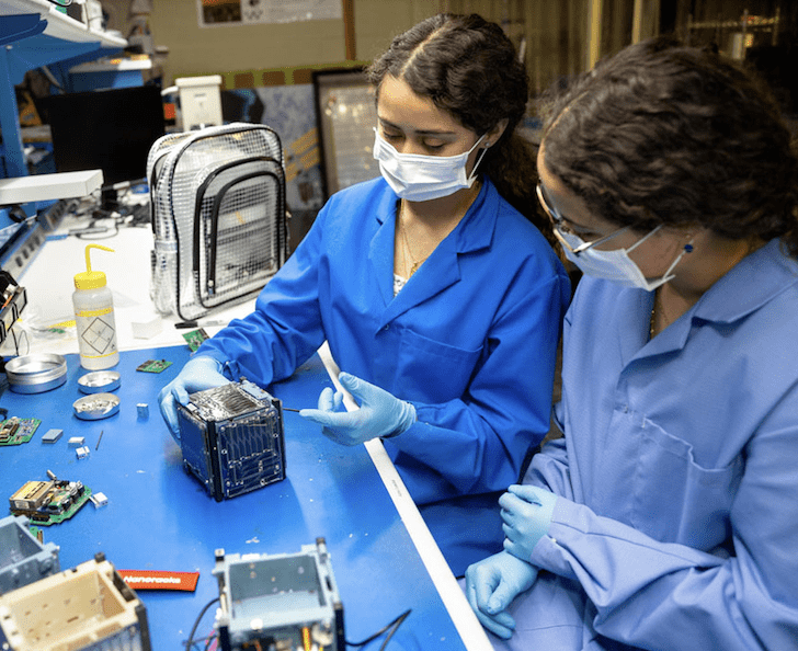 Twin sisters and mechanical engineering majors Verena (L) and Ashley (R) Padres in the Space Systems Design Studio Lab. She is showing Alpha CubeSat (light sail-carrying, cube-shaped satellite)