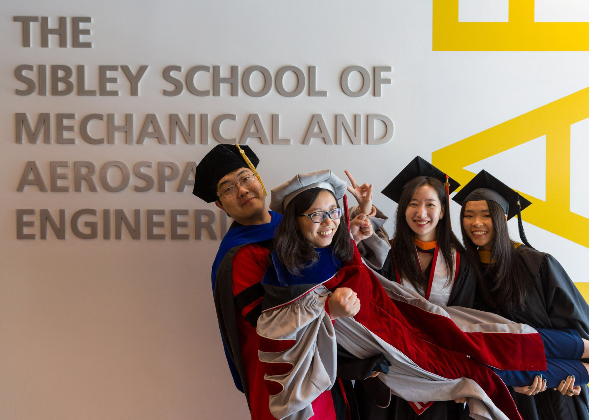 Three students in caps and gowns lift Professor Zhiting Tian, who is also wearing a cap and gown. The group is posing in front of a sign that says 