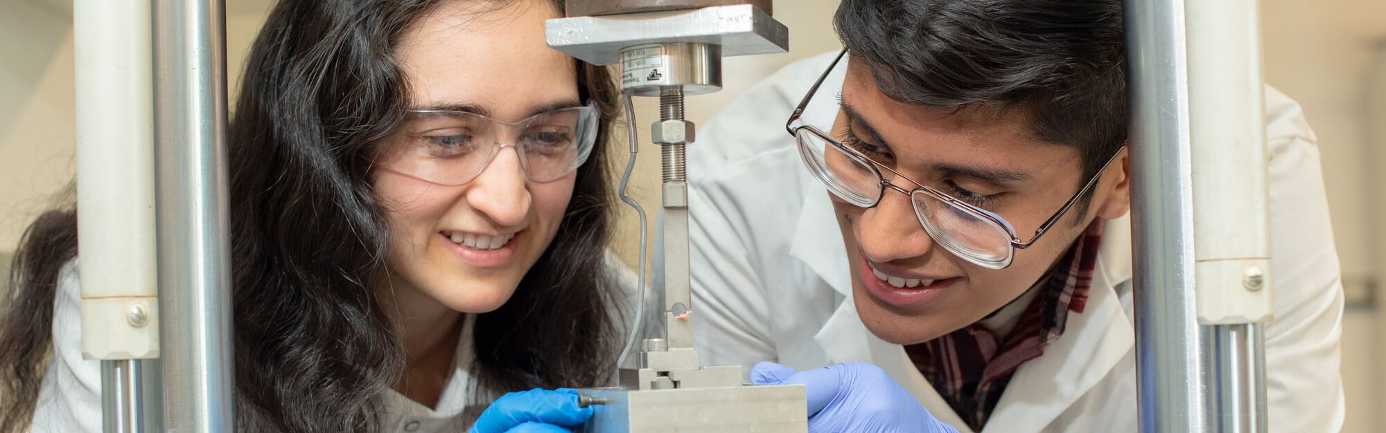 Two students in white lab coats and protective goggles examine a piece of equipment in the Hernandez lab