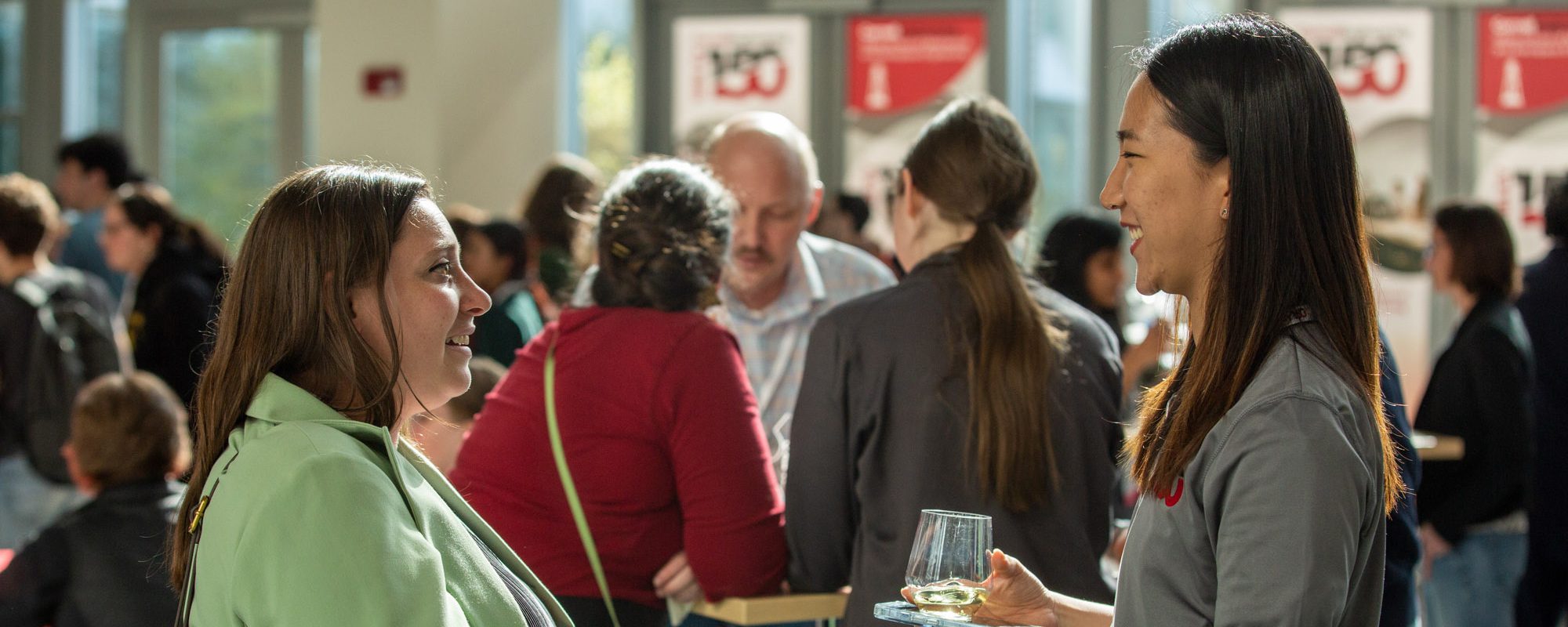 Two people hold drinks and smile at each other amid a crown in Duffield Hall.