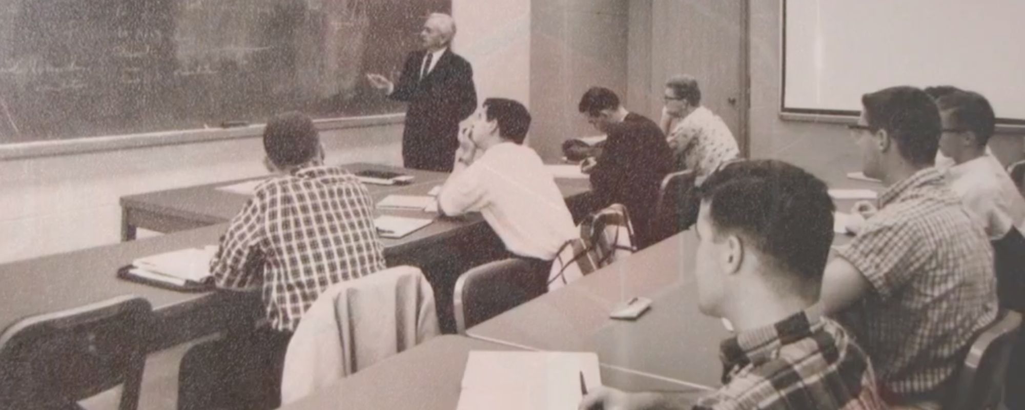 Historic photo of students sitting in rows staring at a professor standing at a large chalk board lecturing in materials science and engineering.