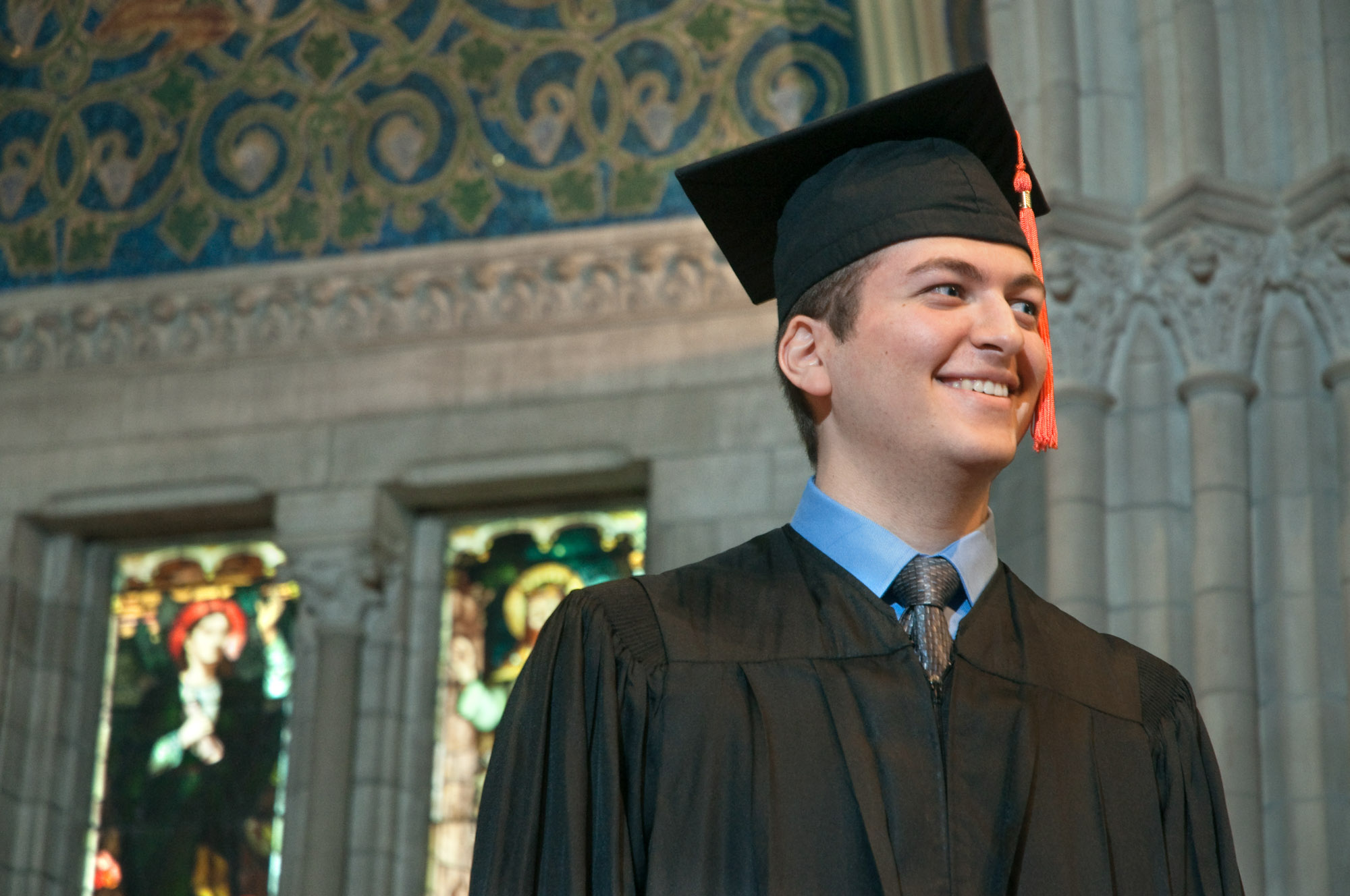 A graduating student stands in Sage Chapel, wearing a black robe and cap.