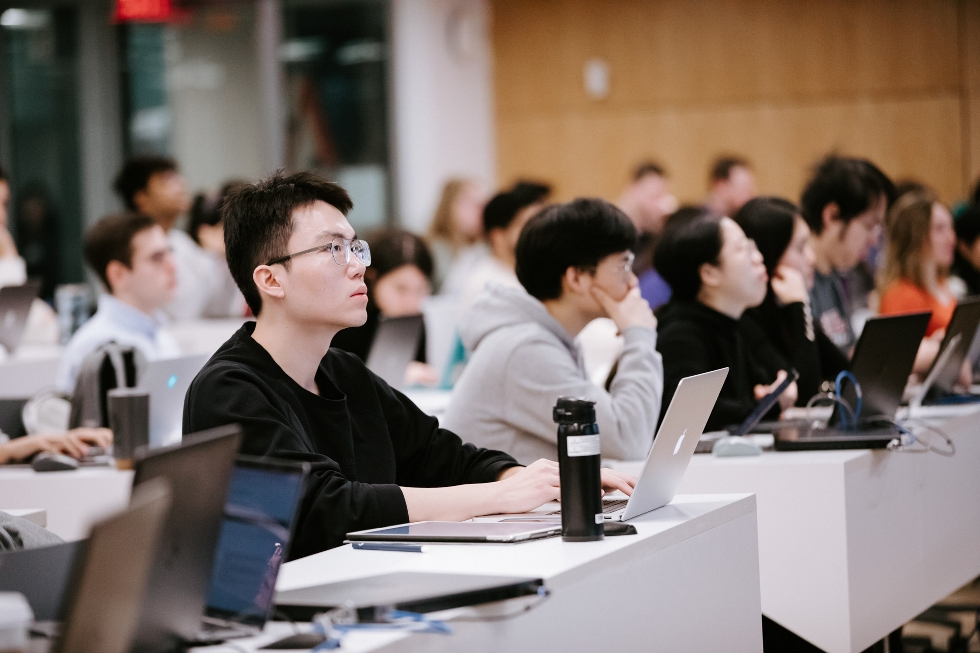 rows of students sit in a lecture hall with laptops open in front of them.