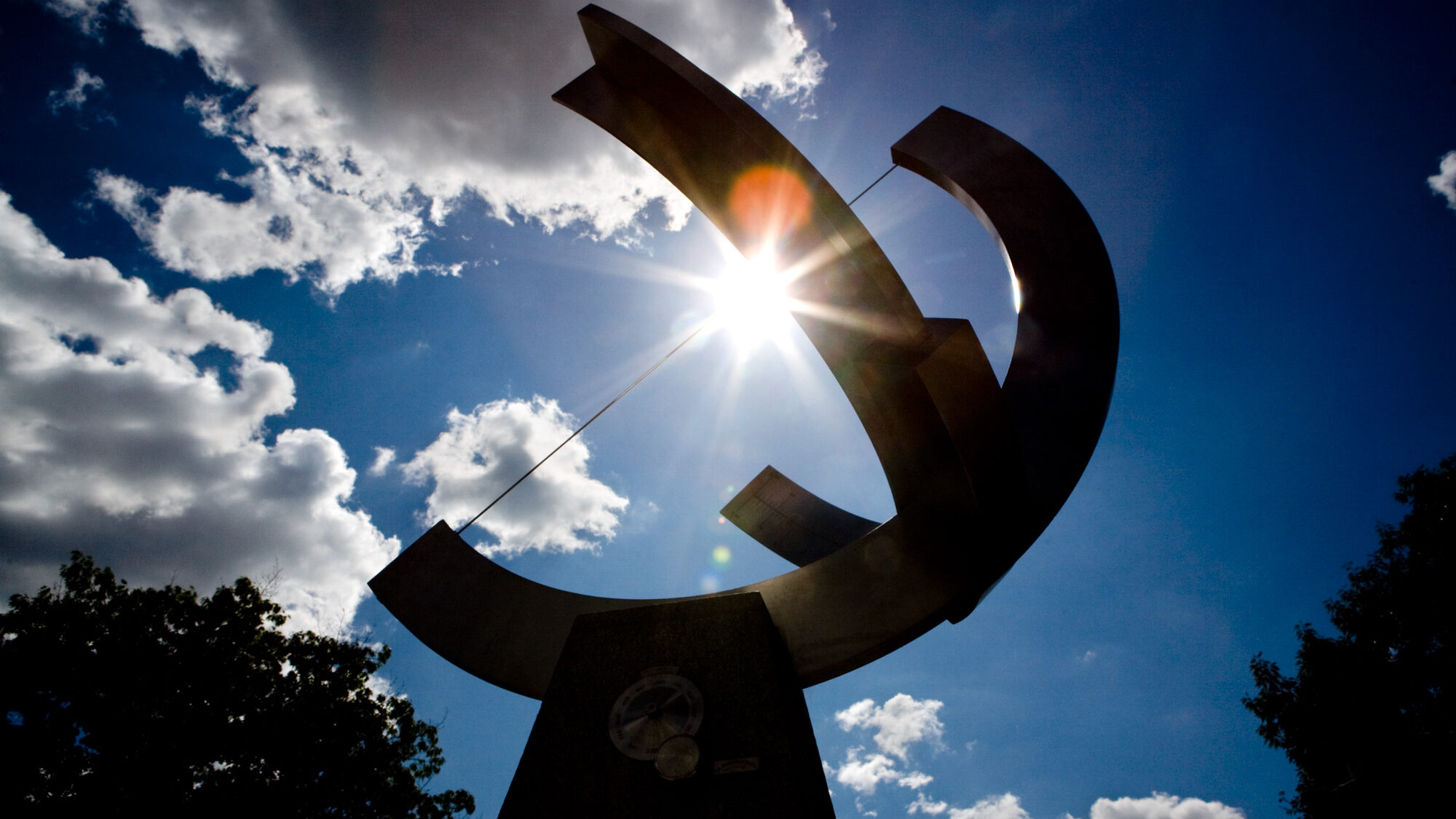 The sun shines through the large matallic sundial on the Pew Engineering Quad. There are clouds in the blue sky above the sundial.