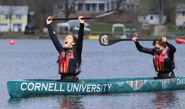 Two women in a concrete canoe hold their paddles up in victory.
