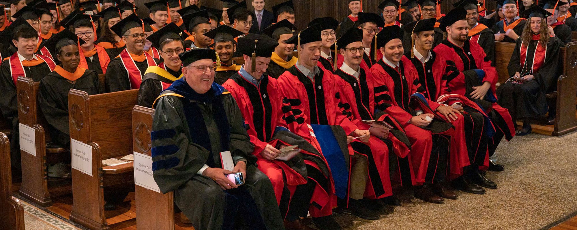 Smiling alumni in graduation robes sit in rows in the Sage Hall benches.