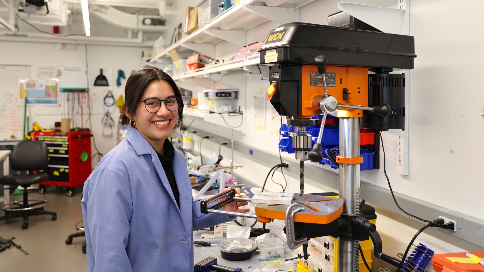 Student wearing lab coat smiles standing by machinery in lab