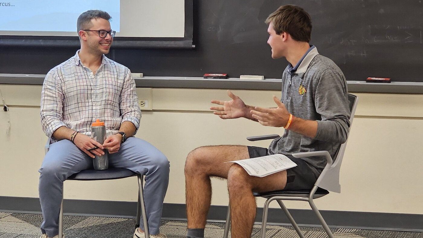 a student and staff member sit on chairs and talk in a classroom