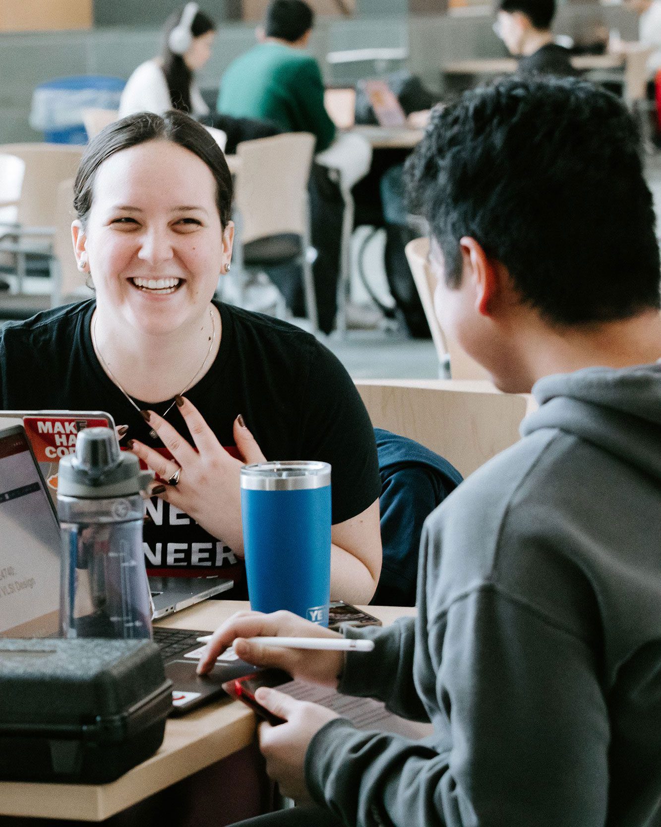 Two students work at their laptops and tablets together at table in Duffield Hall, talking and laughing